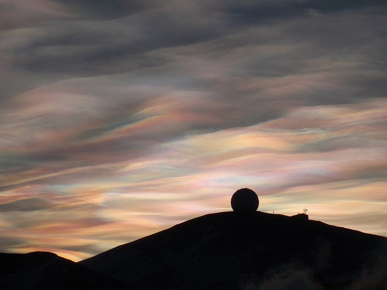 Iridescent nacreous clouds, a rare cloud formation, glow over a dark mountain with a radar dome. Iridescent nacreous clouds, a rare cloud formation, glow over a dark mountain with a radar dome.