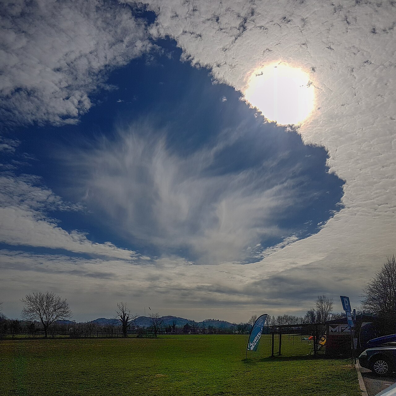 A large, circular hole punch cloud with wispy fallstreaks in a blue sky, sunlight shining through. A large, circular hole punch cloud with wispy fallstreaks in a blue sky, sunlight shining through.