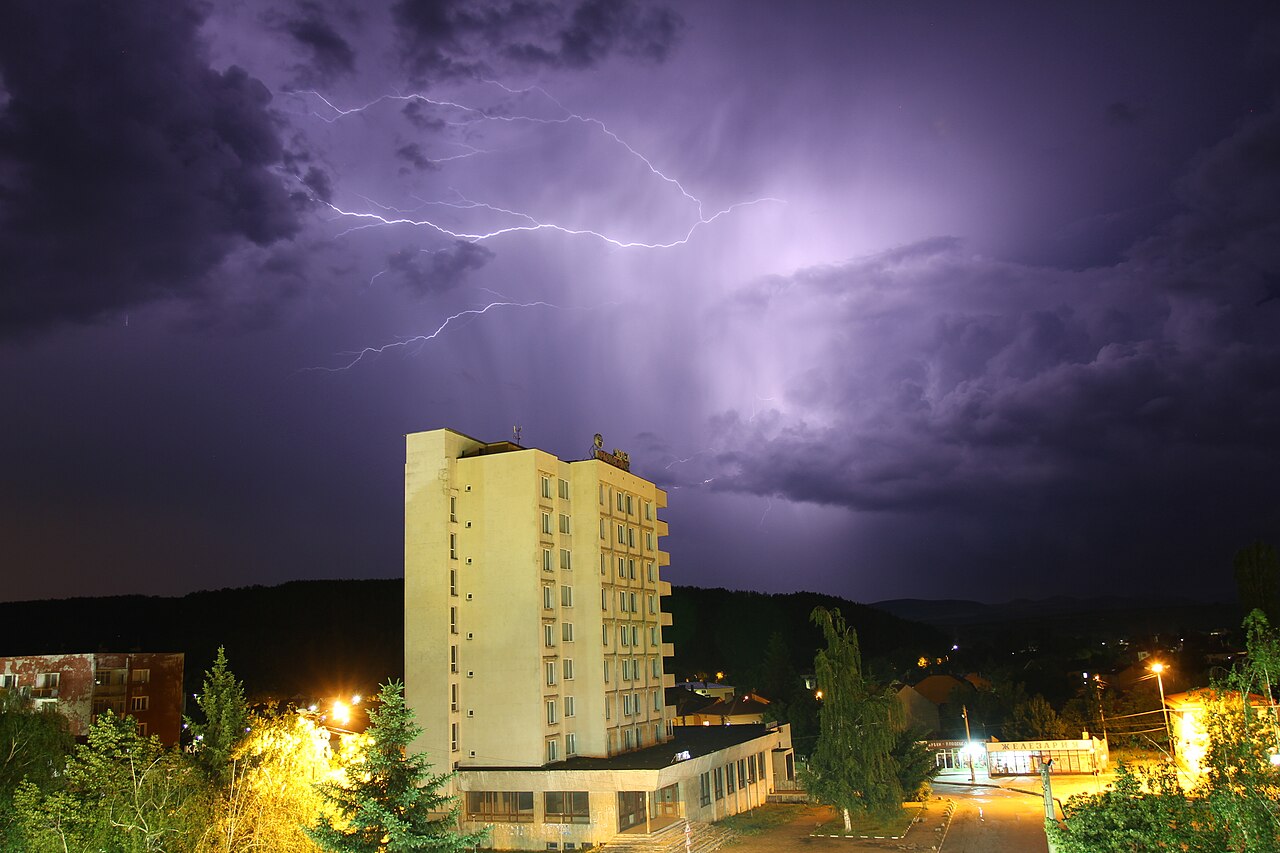 Bright cloud-to-cloud lightning illuminates a dark, stormy sky above a multistory building and town at night. Bright cloud-to-cloud lightning illuminates a dark, stormy sky above a multistory building and town at night.