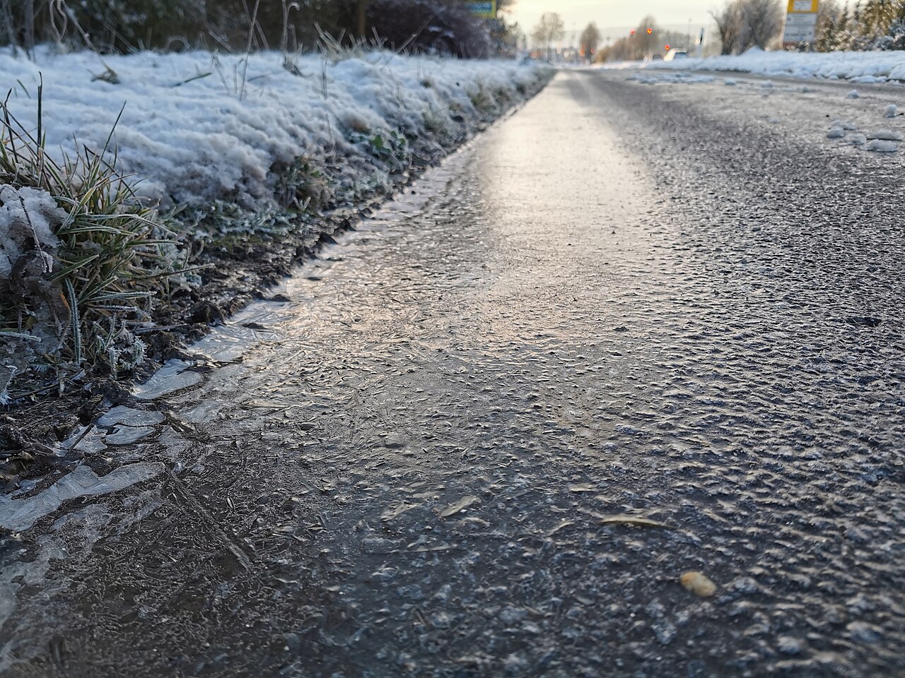 Glistening black ice on a road next to a snow-dusted verge, a hidden hazard for drivers.