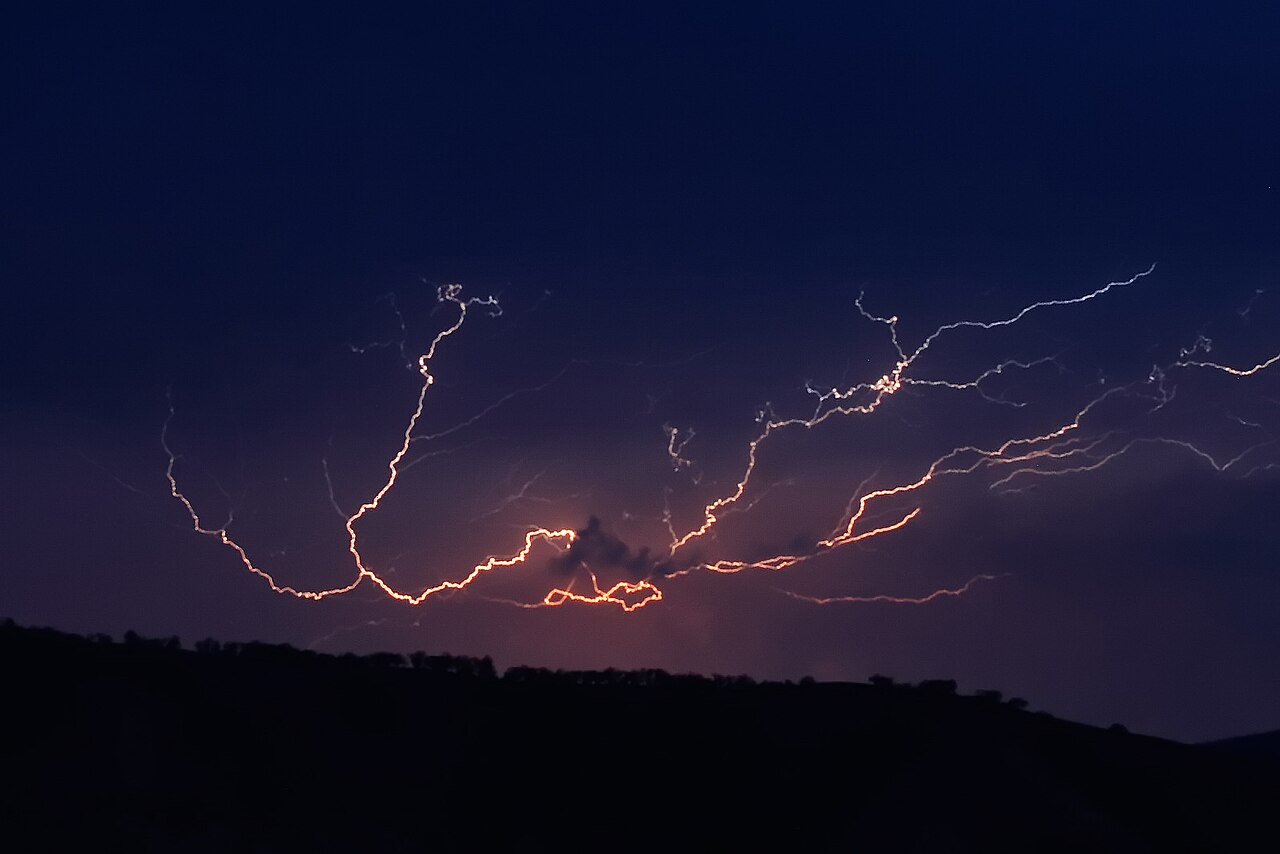 Intricate orange cloud-to-cloud lightning illuminates a dark night sky above a silhouetted horizon, showcasing atmospheric el Intricate orange cloud-to-cloud lightning illuminates a dark night sky above a silhouetted horizon, showcasing atmospheric el