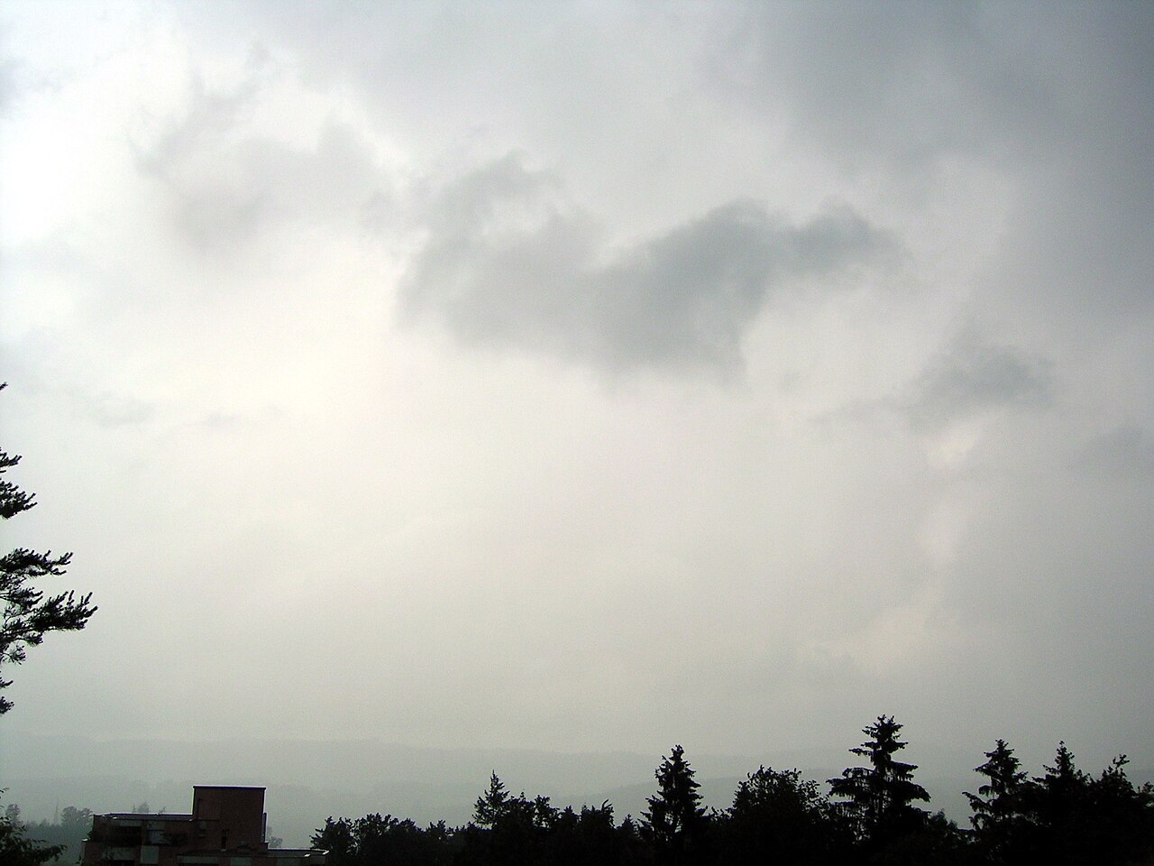 Overcast sky with dark nimbostratus clouds, indicating continuous rain, above a distant tree line and building. Overcast sky with dark nimbostratus clouds, indicating continuous rain, above a distant tree line and building.